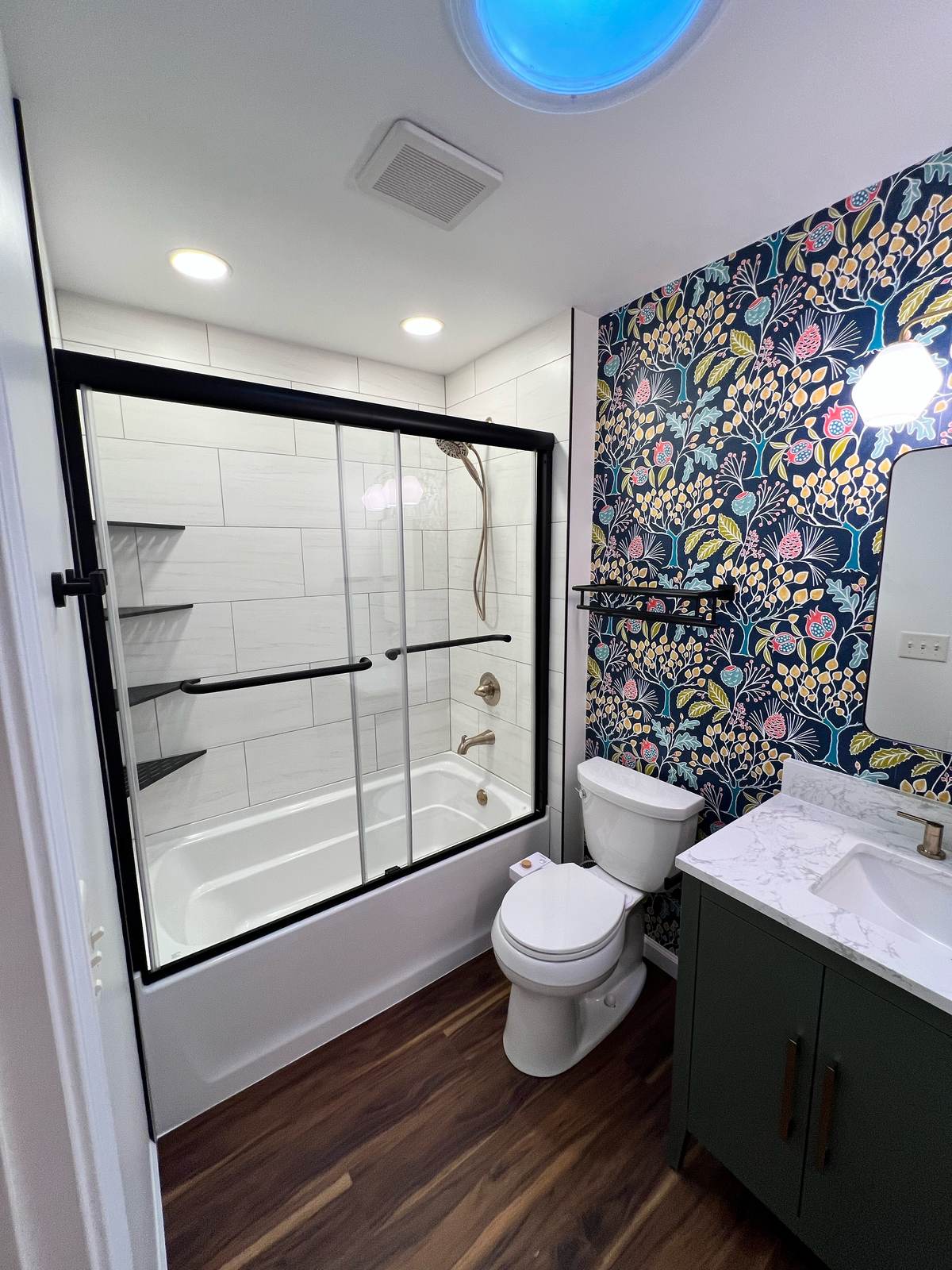 Full bathroom with bold botanical wallpaper, green vanity with marble-look top, black-framed sliding tub door, and wood-look floor