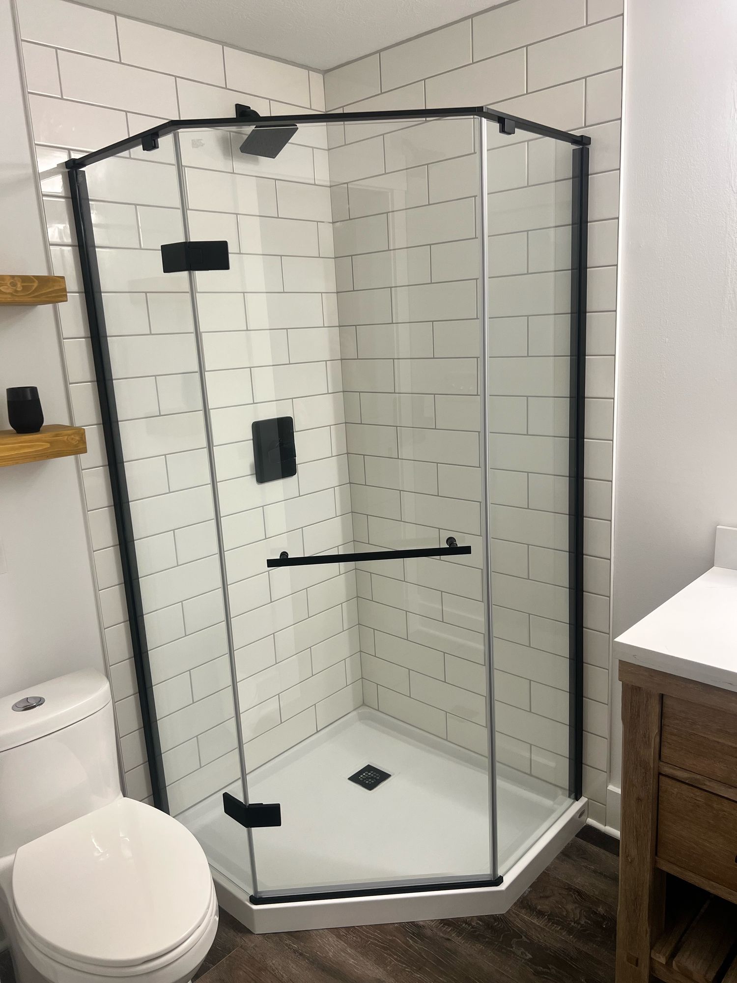 Neo-angle glass shower with a matte black frame, a white base, white subway tile to the ceiling, and a black square rain head and valve, with wood floating shelves and a rustic wood vanity