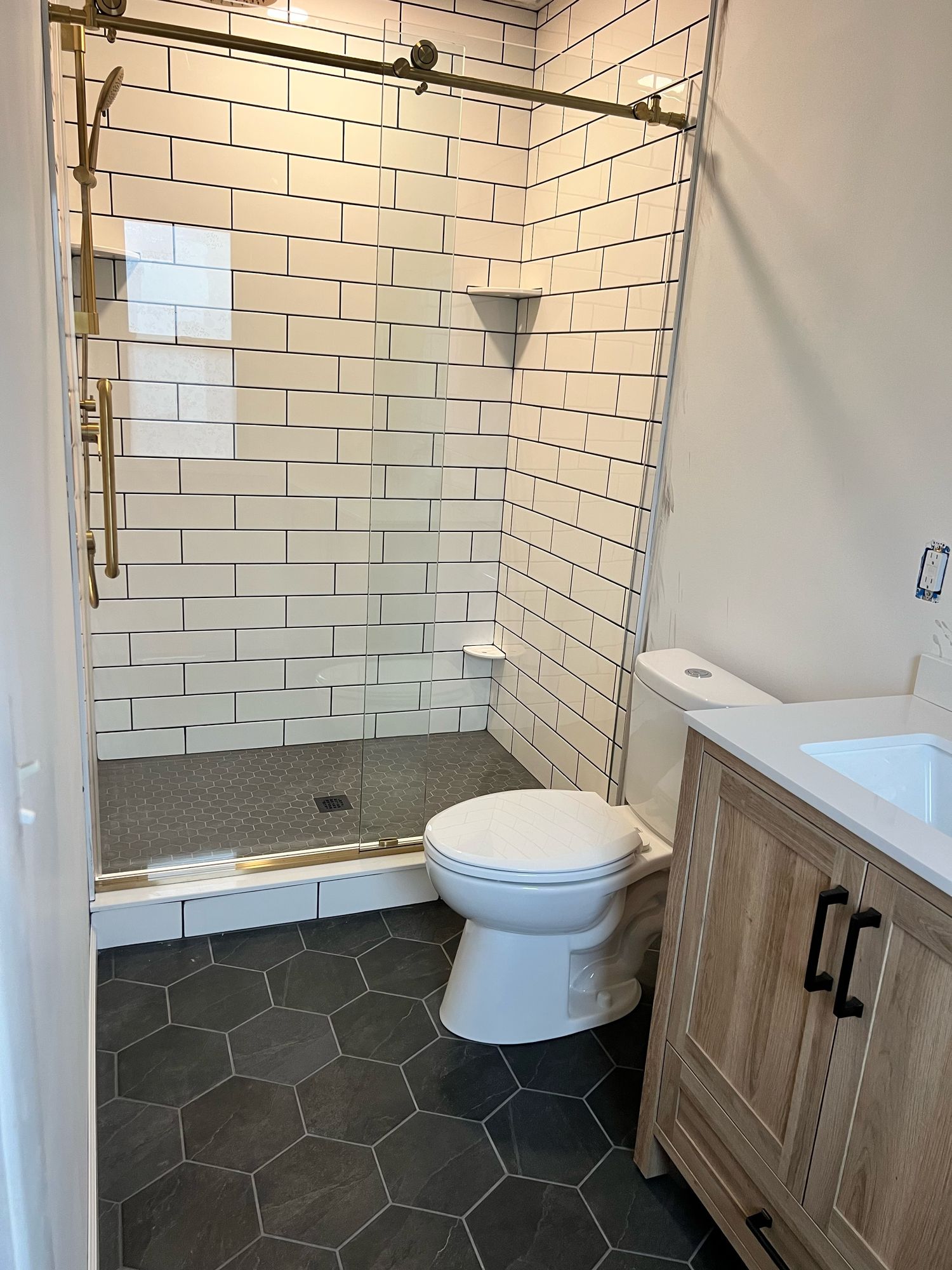 Subway tile walk-in shower with black grout, a brushed gold sliding glass door, and a gray hex mosaic floor