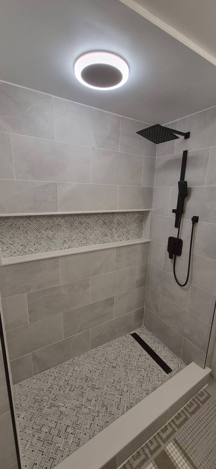 Walk-in shower with large gray porcelain tile, marble herringbone niche and floor, linear drain, and matte black rain shower