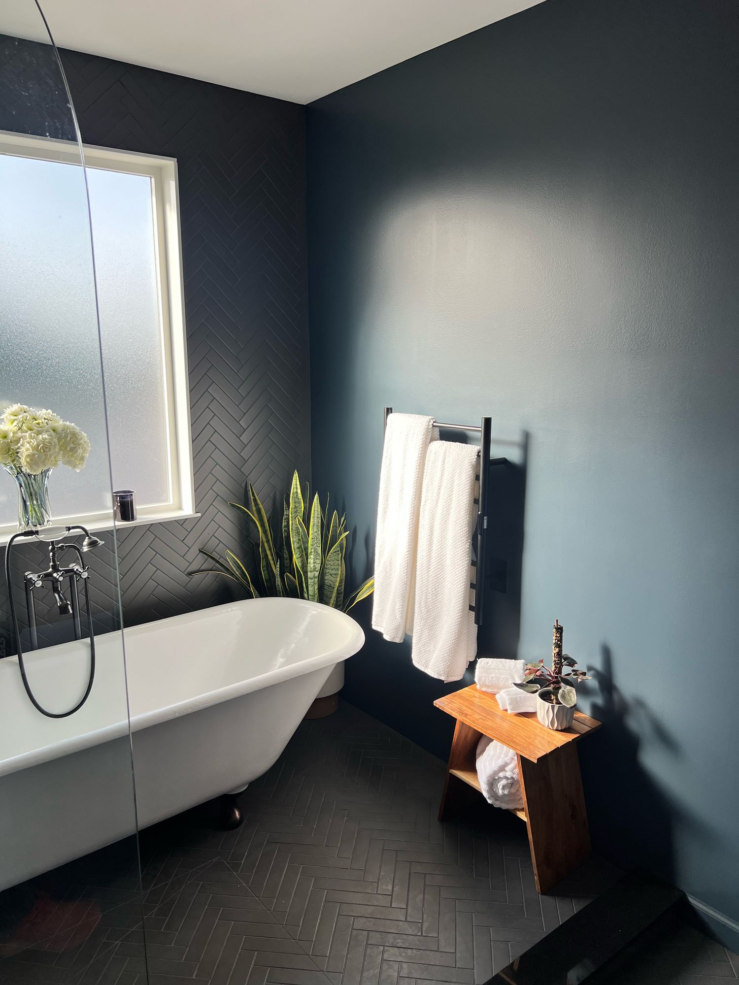 White clawfoot tub with a floor-mount tub filler under a frosted window, a charcoal herringbone accent wall, and a heated towel rack on a dark teal wall