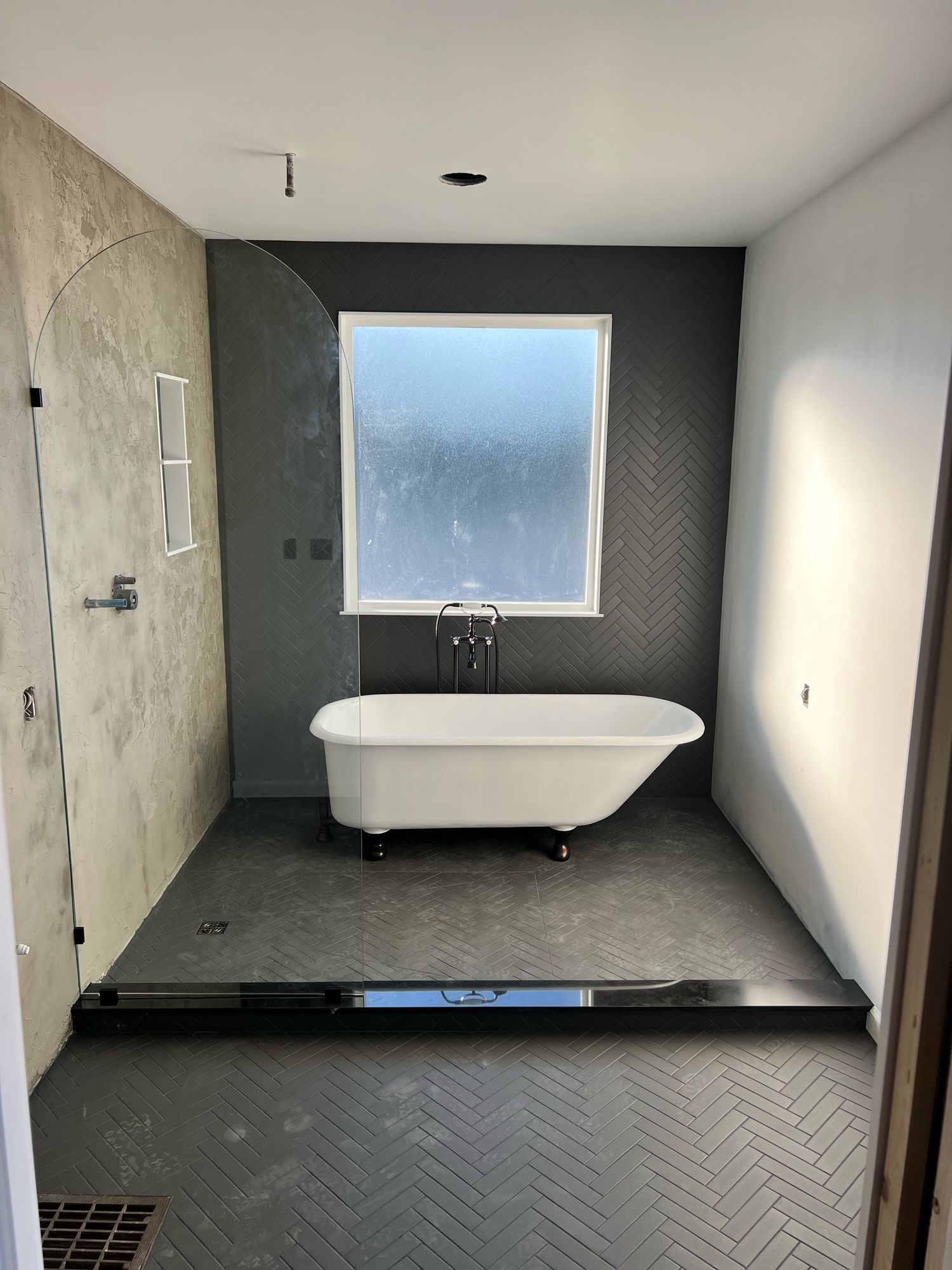 Bathroom before trim-out, with a white clawfoot tub and floor-mount filler under a frosted window, a charcoal herringbone accent wall and floor, a black curb, and a curved glass shower panel