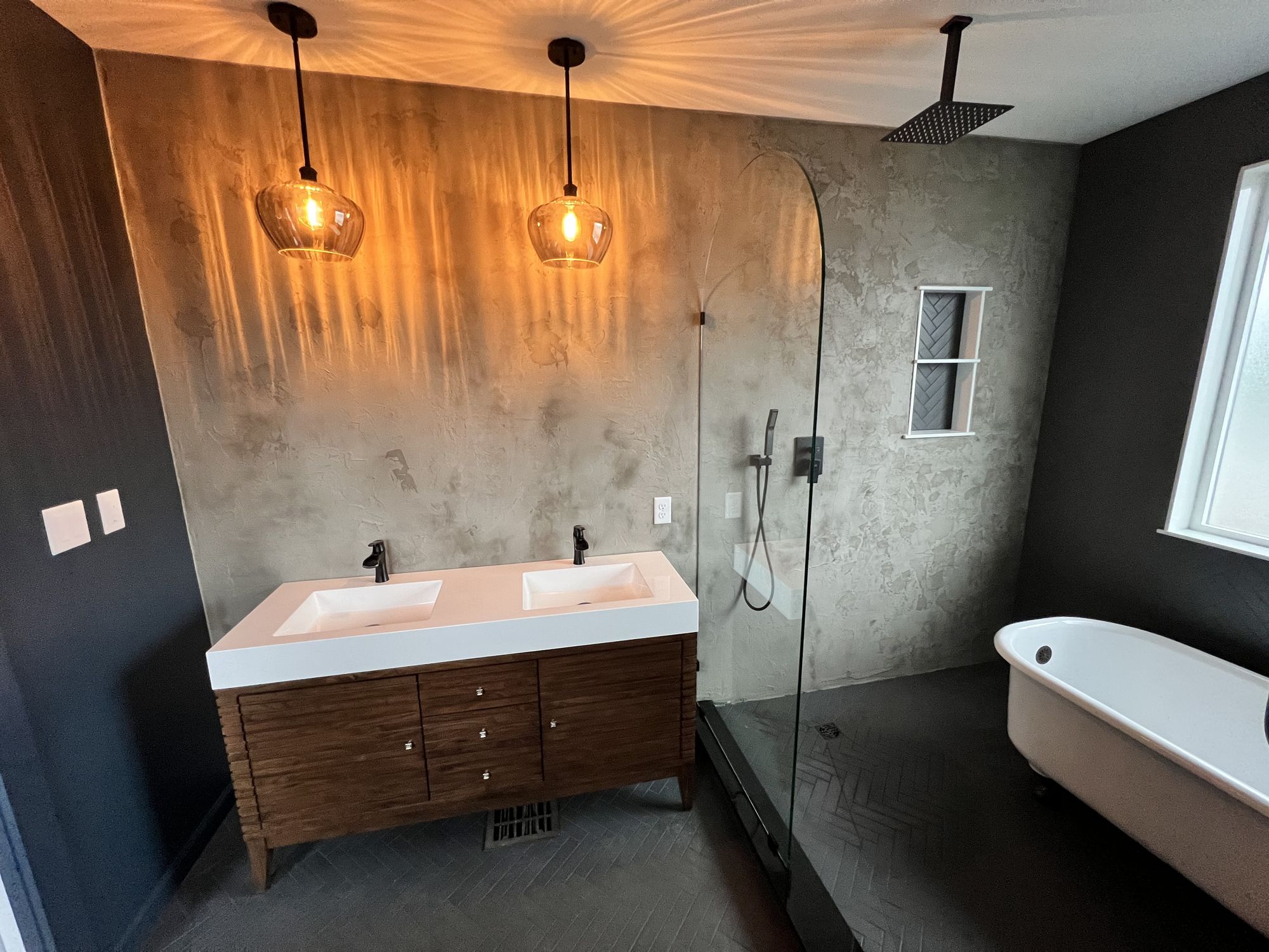 Bathroom with textured gray plaster walls, a walnut slatted double vanity with a white double-sink top, two amber glass pendants, and a curved frameless glass walk-in shower