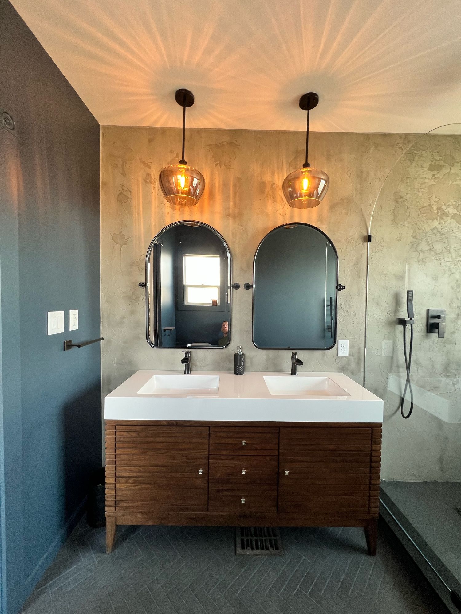 Walnut slatted double vanity with a white integrated double-sink top, matte black faucets, two arched black mirrors, and two amber glass pendant lights