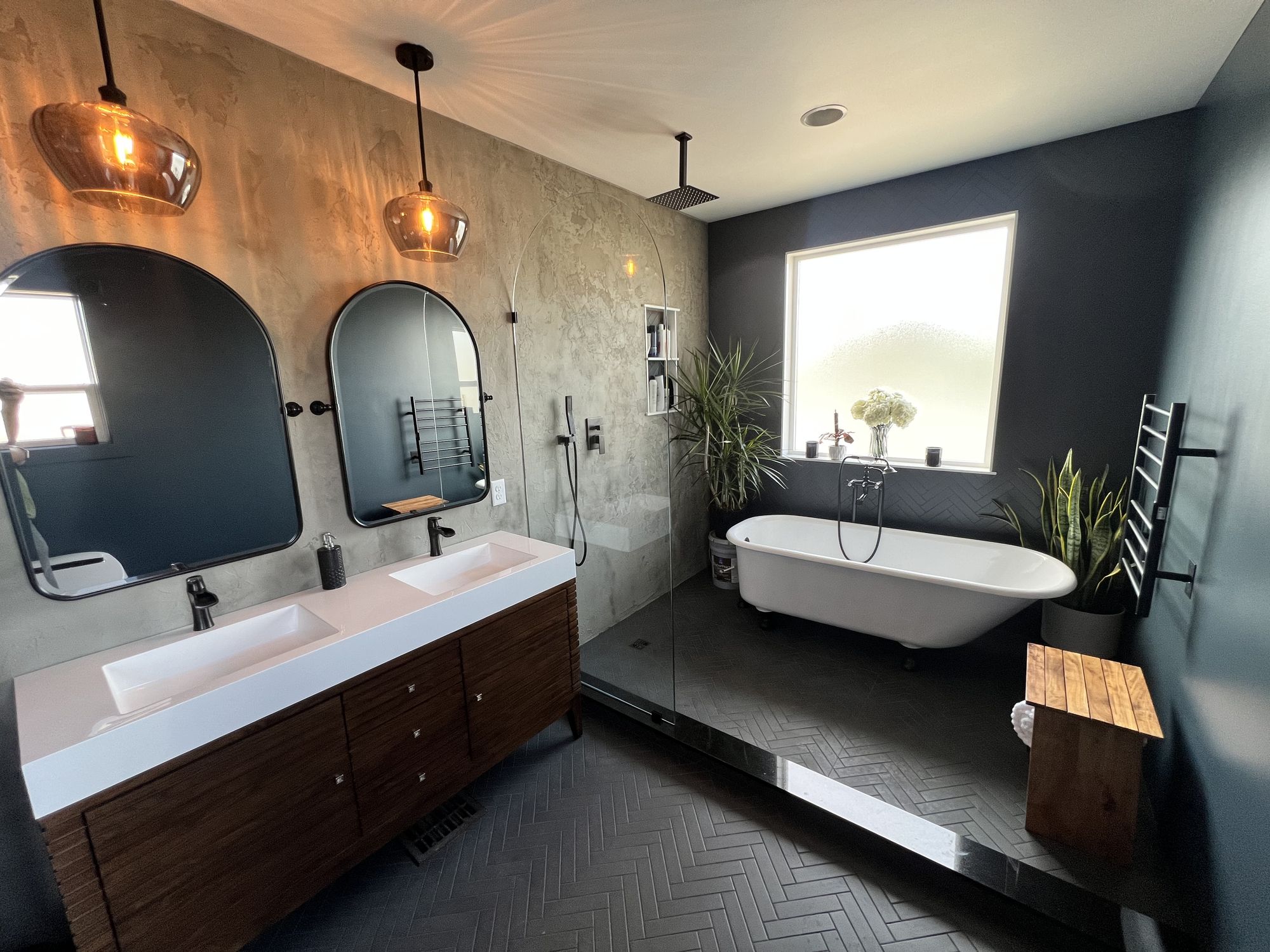 Bathroom with a walnut double vanity, arched black mirrors, a curved glass walk-in shower, and a white clawfoot tub behind a black curb on charcoal herringbone tile