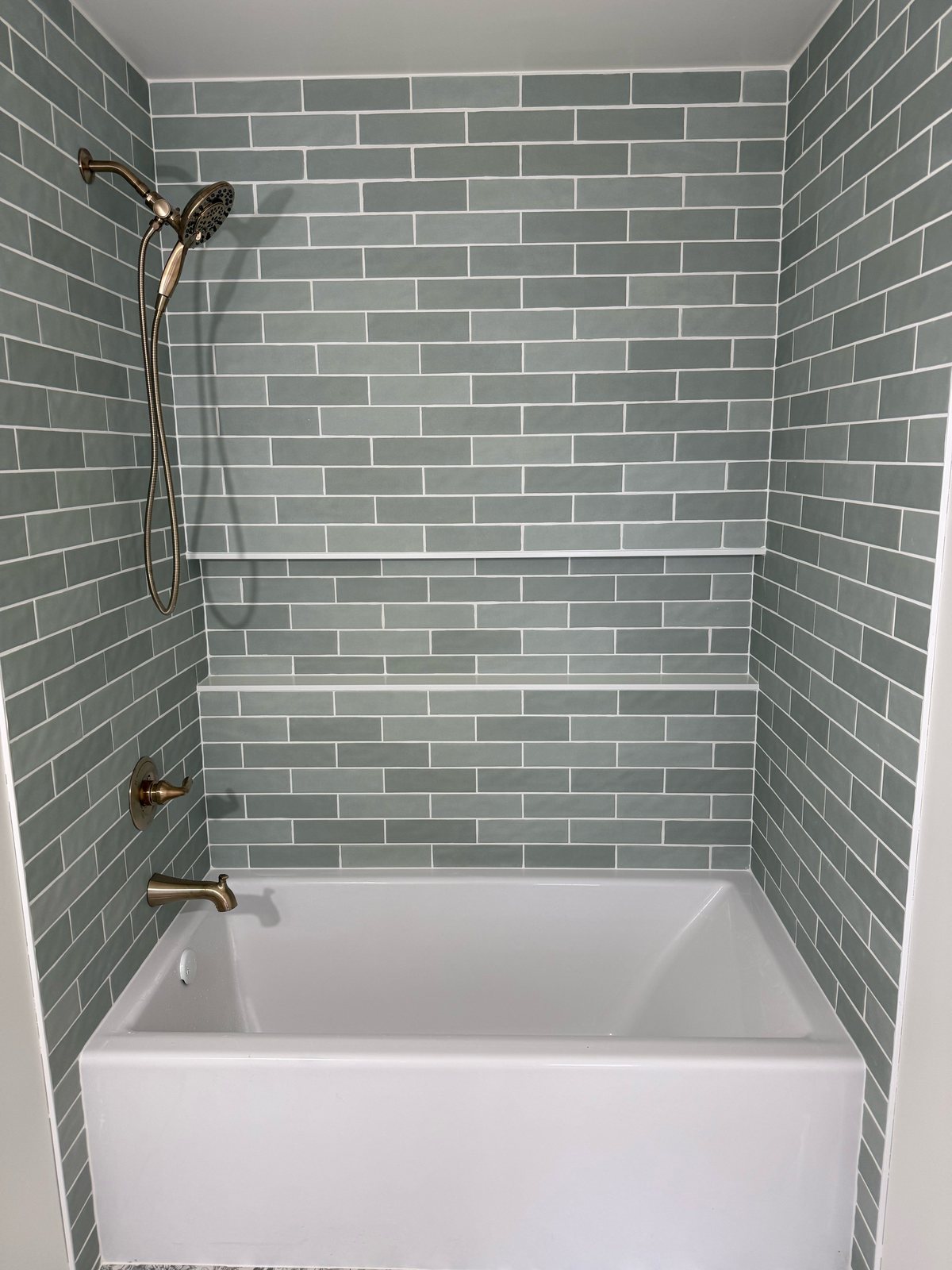 Tub-shower with sage green subway tile, tiled shelves, white alcove tub, and champagne bronze handheld shower