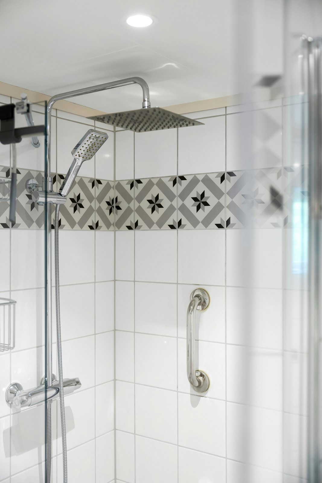 White tile shower with a stainless grab bar, handheld shower, and patterned tile band